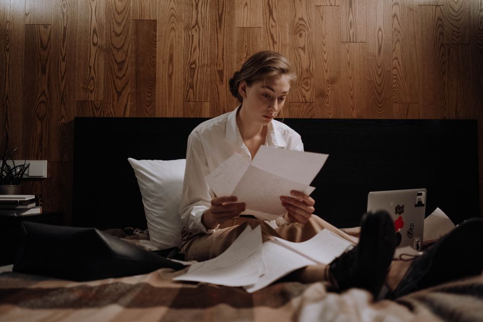 Woman sitting on bed, working from home with laptop and paperwork, focused and thoughtful.