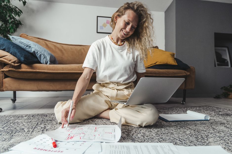 Smiling woman sitting on floor, working on laptop, surrounded by papers in cozy home setting.