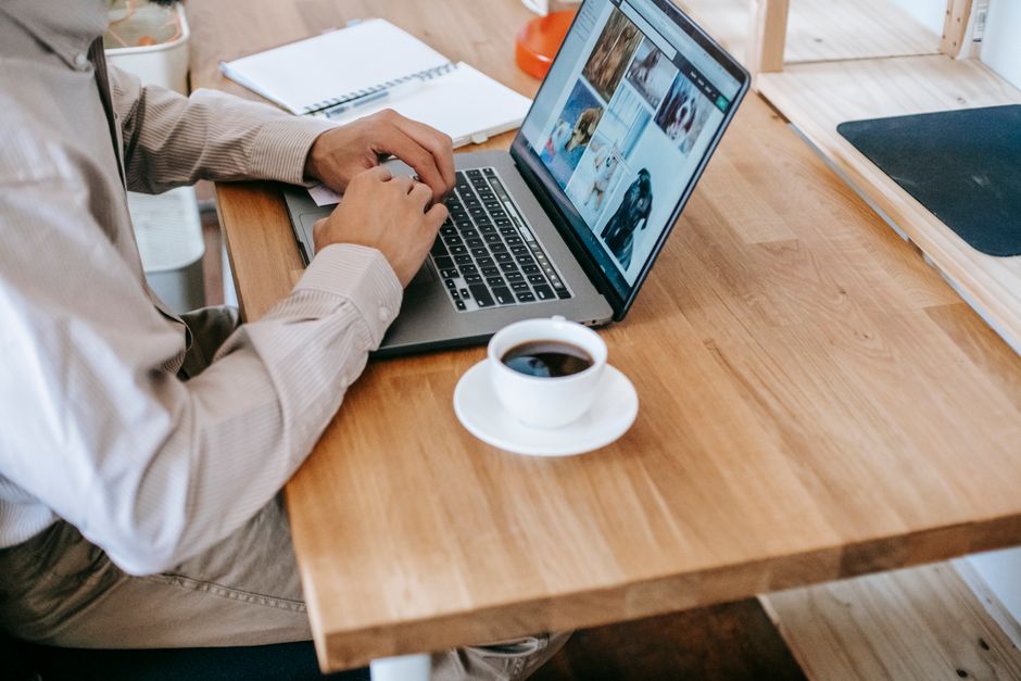 High angle of crop unrecognizable man freelancer in casual outfit sitting at table and working on computer while drinking coffee near notebook in light workplace