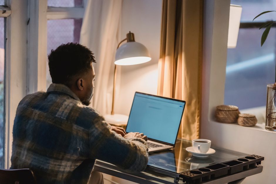Back view of concentrated African American male typing on keyboard of laptop while sitting at table with cup of coffee and light from lamp